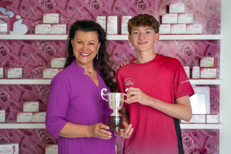„Prof. Anita Frauwallner vom Institut Allergosan und der junge Athlet Moritz Freitag halten gemeinsam einen silbernen Pokal vor einem Regal mit OMNi-BiOTiC®-Produkten. Beide lächeln in die Kamera.“