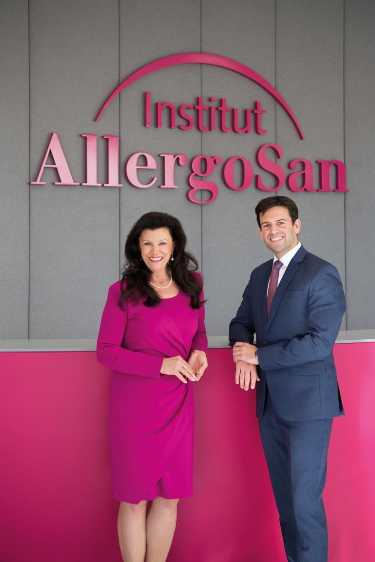 Zwei Personen, Anita Frauwallner im pinken Kleid und Bernd Assinger im Anzug, stehen lächelnd vor dem Logo des Instituts AllergoSan.