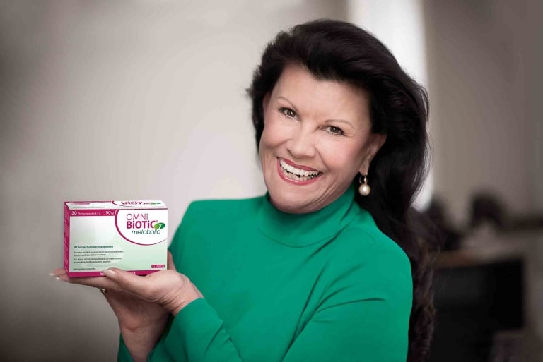 Prof. Anita Frauwallner in einem smaragdgrünen Oberteil hält lächelnd eine Packung „OMNi-BiOTiC® metabolic“ in die Kamera