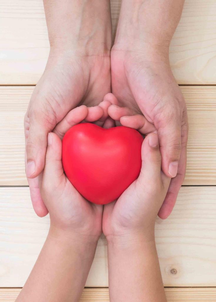 Kinderhände halten ein rotes Herz, umschlossen von schützenden Erwachsenen-Händen.