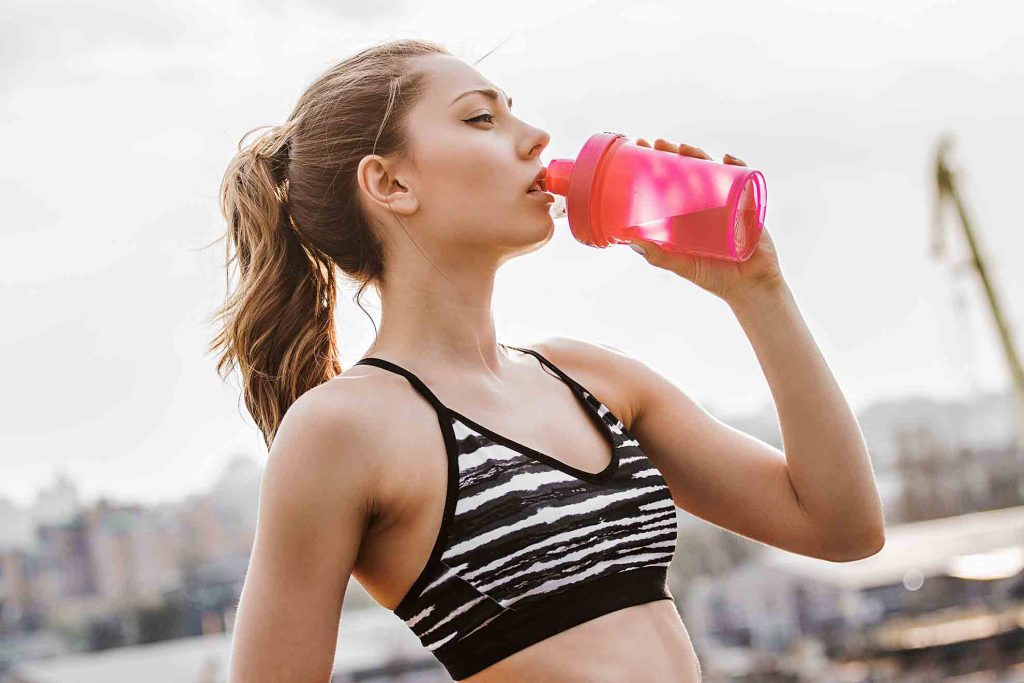 Junge sportliche Frau trinkt nach dem Training aus einer pinken Trinkflasche – Symbol für Fitness, Hydration und gesundes Körpergefühl